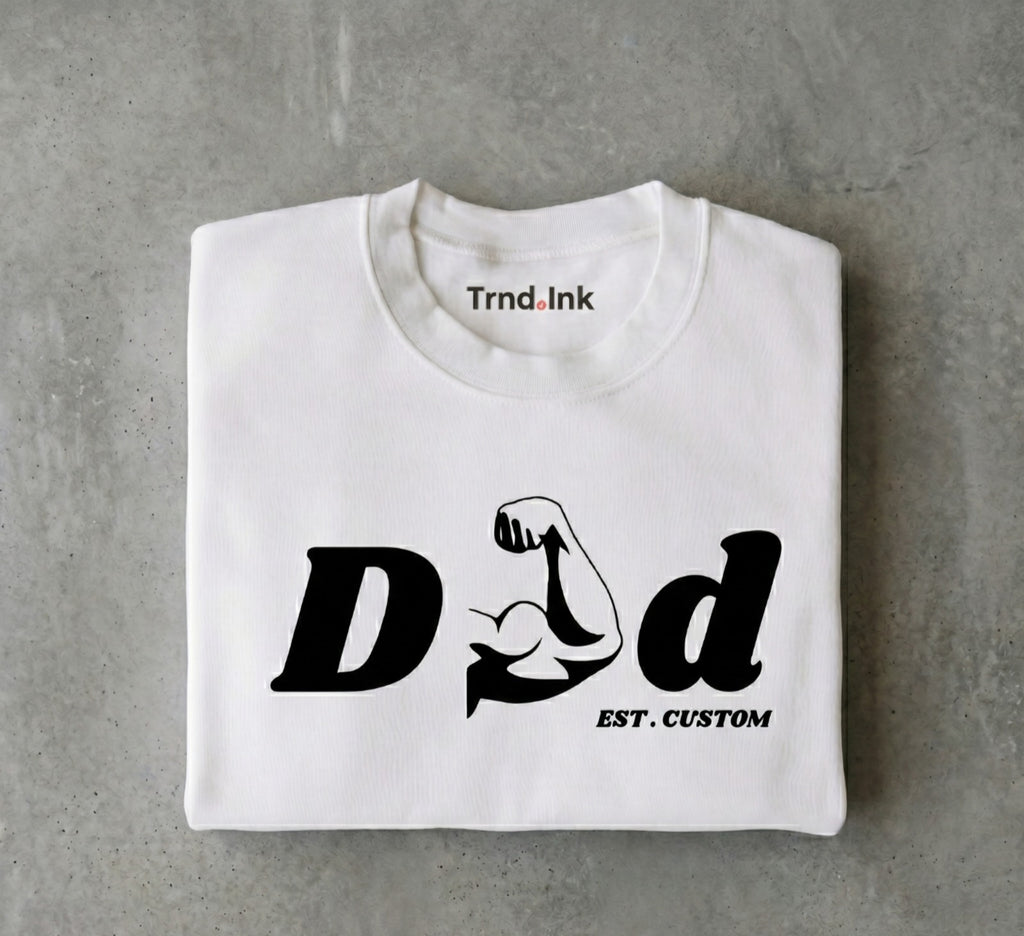 Fotografía de producto estilo flat lay de una camiseta blanca cuidadosamente doblada sobre una superficie gris texturizada con efecto de concreto. En el frente de la prenda destaca el diseño minimalista en tinta negra "DAD", donde la letra 'A' es reemplazada por un brazo musculoso flexionando el bíceps, con la frase "EST. CUSTOM" debajo. El logo de Trnd.Ink se aprecia nítidamente en la parte superior central interna del cuello.