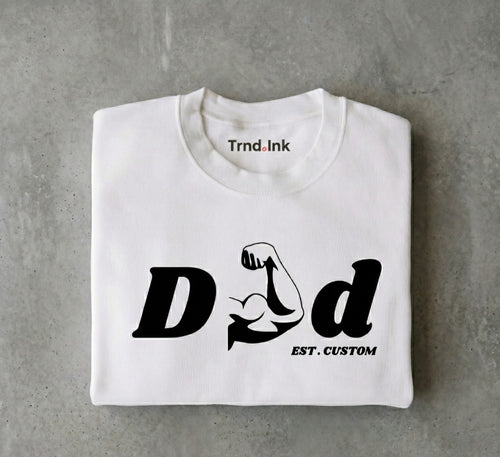 Fotografía de producto estilo flat lay de una camiseta blanca cuidadosamente doblada sobre una superficie gris texturizada con efecto de concreto. En el frente de la prenda destaca el diseño minimalista en tinta negra "DAD", donde la letra 'A' es reemplazada por un brazo musculoso flexionando el bíceps, con la frase "EST. CUSTOM" debajo. El logo de Trnd.Ink se aprecia nítidamente en la parte superior central interna del cuello.