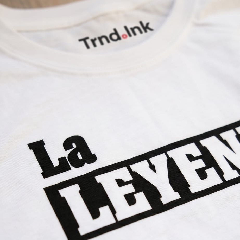 Primer plano de detalle del cuello redondo de una camiseta blanca de la marca Trnd.Ink. Se observa el logo de Trnd.Ink sin variaciones impreso en la parte superior central interna del cuello y el inicio de la tipografía negra del diseño "La Leyenda" estampado en el pecho.