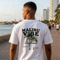 Hombre caminando junto al mar luciendo una camiseta blanca con diseño gráfico de palmeras Malibu Vibe y logo de Trnd.ink en el pecho
