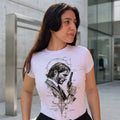 Mujer joven modelando una camiseta blanca con ilustración artística frontal de John Wick en un entorno urbano con arquitectura moderna de fondo.