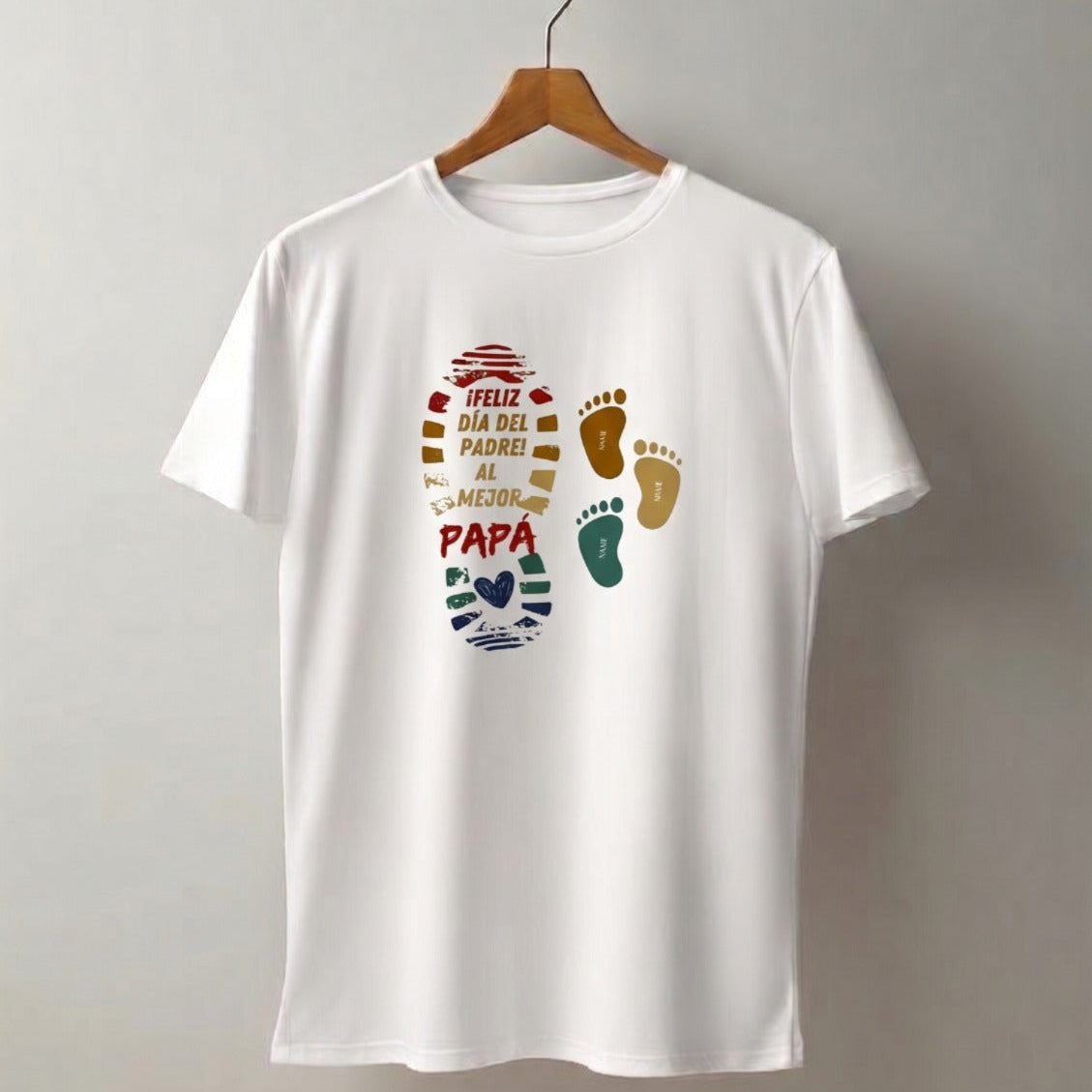 otografía de estudio de una camiseta blanca de cuello redondo para hombre, colgada en un gancho de madera sobre una pared gris neutra. La prenda de la marca Trnd.Ink exhibe un estampado central con una huella grande y colorida con la frase "¡FELIZ DÍA DEL PADRE! AL MEJOR PAPÁ", acompañada de dos pequeñas huellas infantiles a un lado.