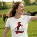 Mujer joven sonriendo al aire libre en un campo con caballos de fondo, vistiendo una camiseta blanca de manga corta de trnd.ink con un estampado frontal de un caballo rojo encabritado.