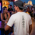 Hombre de espalda usando una camiseta blanca con estampado vertical de fases lunares en una terraza nocturna, estilo de vida urbano y moderno.