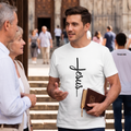 Hombre joven vistiendo una camiseta blanca de cuello redondo con un diseño minimalista de la palabra "Jesus" formando una cruz negra en el pecho. Sostiene una Biblia bajo el brazo mientras conversa en un entorno exterior iluminado.