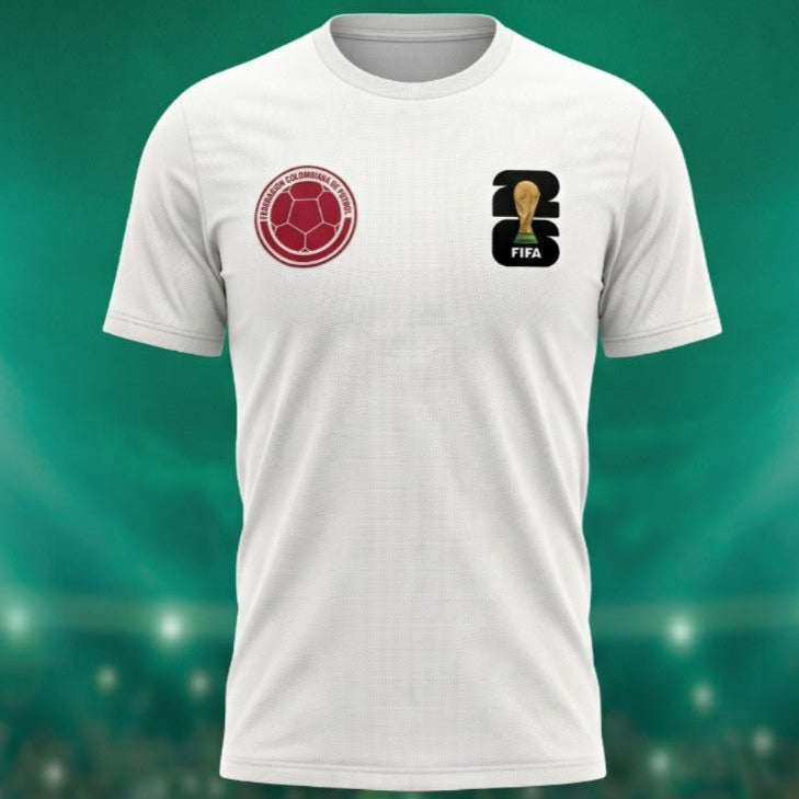 Vista frontal de un mockup de camiseta de fútbol de color blanco perteneciente a la colección de la marca Trnd.Ink. La prenda presenta en el lado derecho del pecho un escudo circular en tono rojo quemado inspirado en la Selección Colombia, mientras que en el lado izquierdo exhibe el logotipo oficial de la Copa Mundial de la FIFA 2026. La camiseta está dispuesta sobre un fondo verde difuminado que simula la atmósfera de un estadio de fútbol iluminado.
