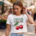 Modelo usando el crop top blanco 'Fresh Cherry' de trnd.ink combinado con jeans, mostrando el diseño de cerezas ilustradas en un ambiente urbano iluminado.