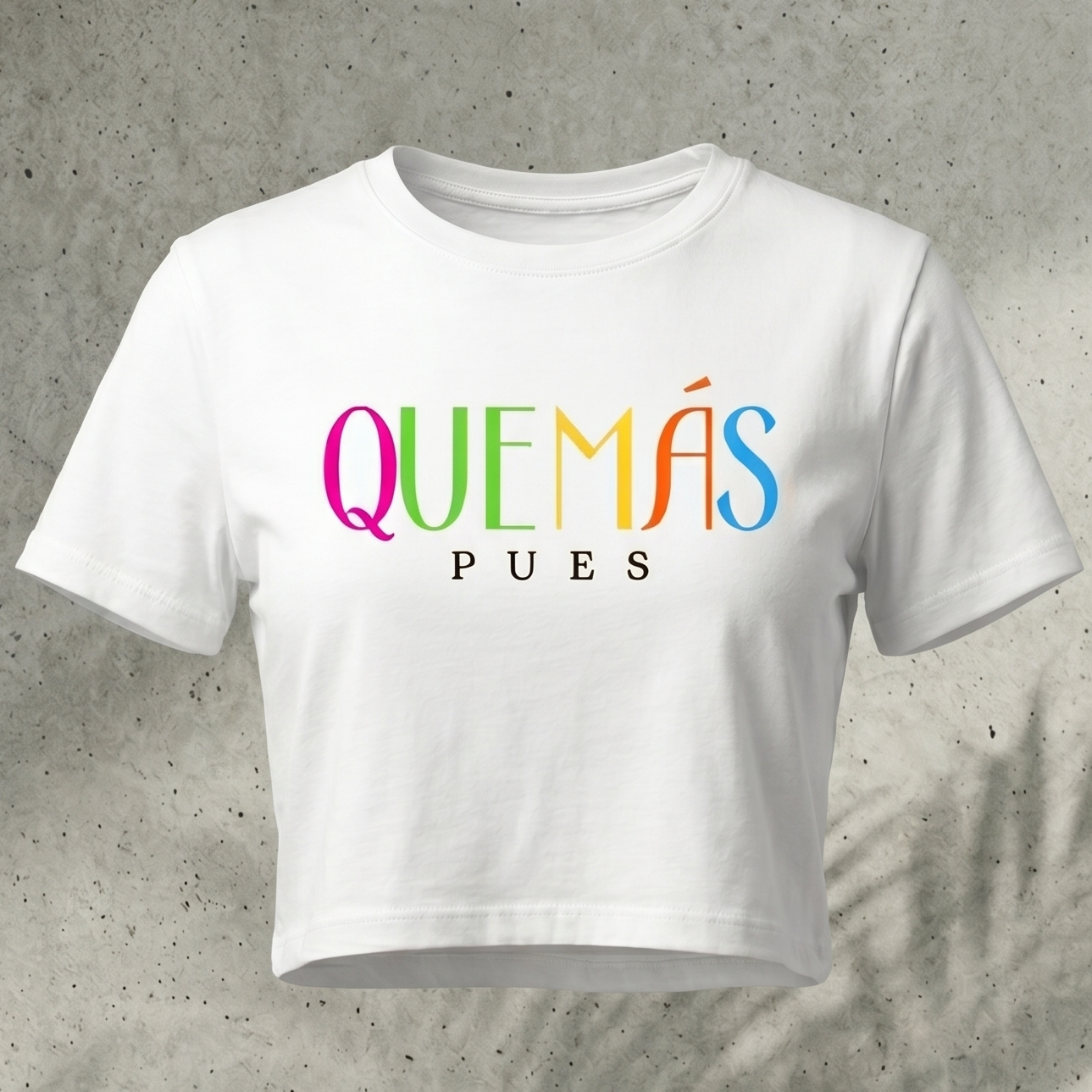 Crop top blanco para mujer con la frase "Qué Más Pues" en letras grandes de colores brillantes y tipografía moderna sobre fondo de concreto.