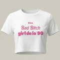 Camiseta blanca tipo crop top para mujer con estampado rosa que dice 'Una Bad Bitch Girl de lo' 90' sobre fondo neutro, estilo moda urbana retro.