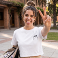 Mujer joven sonriendo y haciendo el signo de la paz, vistiendo una camiseta crop top blanca con un pequeño estampado negro de un personaje minimalista con gafas. Se encuentra en un entorno exterior soleado cargando una mochila con pines.