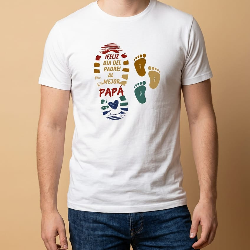 Mockup de un hombre vistiendo una camiseta blanca de manga corta con un diseño estampado para el Día del Padre de la marca Trnd.Ink. El gráfico central muestra la silueta de una huella de pie grande rellena con la frase "¡FELIZ DÍA DEL PADRE! AL MEJOR PAPÁ" en letras de colores, acompañada al lado por dos huellas pequeñas infantiles en tonos ocre y verde.