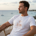 Foto lifestyle de un modelo masculino vistiendo la camiseta blanca "Definitivamente Sí Debí" de Trnd.ink con estampado terracota, sentado en una silla de mimbre mirando a una bahía costera al atardecer.