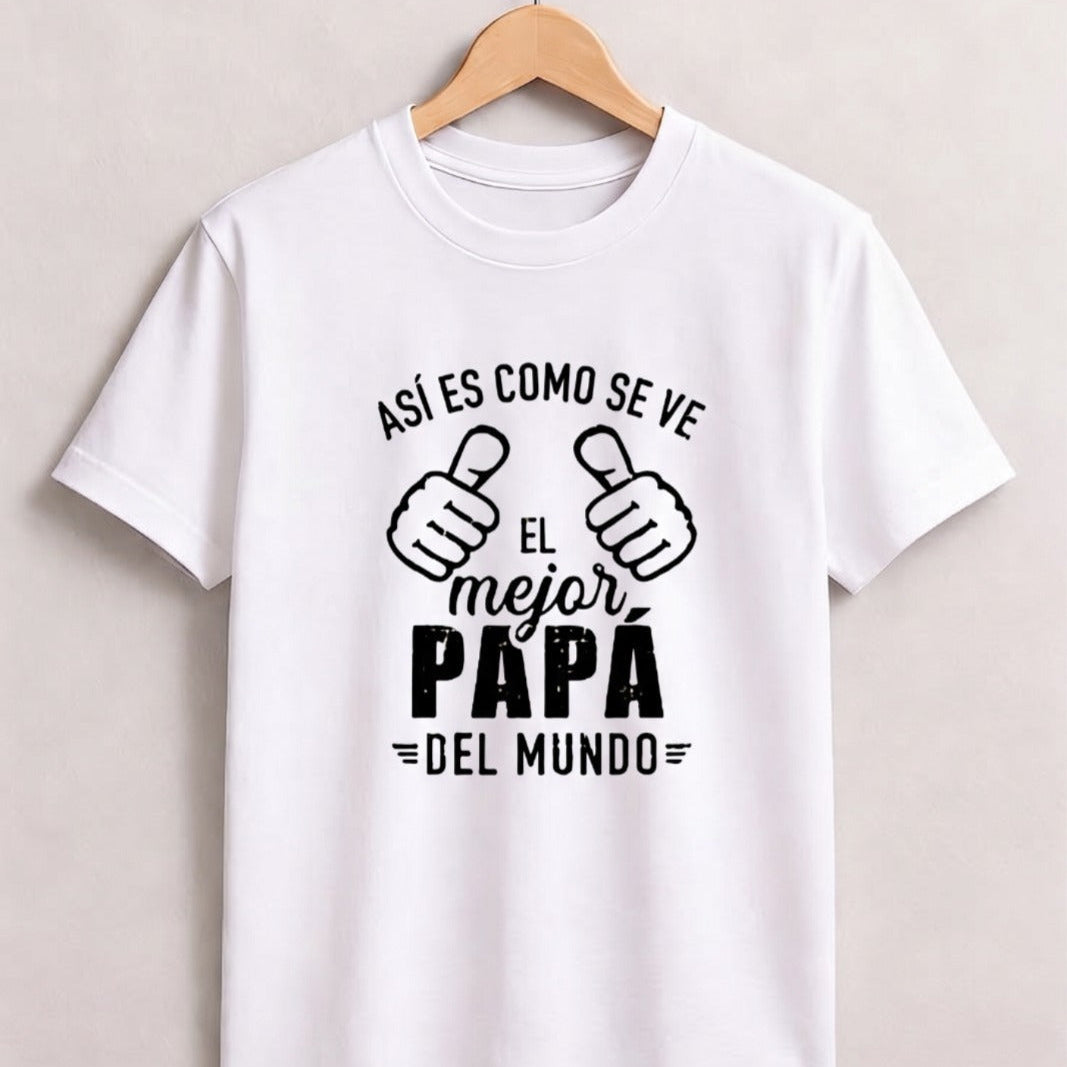 Fotografía de catálogo de una camiseta blanca de cuello redondo para hombre, colgada en un gancho de madera sobre un fondo plano de tono beige claro. La prenda de la marca Trnd.Ink exhibe un estampado central en tinta negra con la frase de estilo minimalista y sarcástico "ASÍ ES COMO SE VE EL mejor PAPÁ =DEL MUNDO=", acompañado de dos manos ilustradas apuntando con los pulgares hacia arriba.