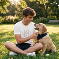 Hombre joven sentado en el césped de un parque jugando con un perro Golden Retriever. El hombre viste una camiseta blanca de Trnd.Ink con un estampado minimalista de una huella de perro y un corazón. Escena soleada de estilo de vida al aire libre con fondo de árboles desenfocados.