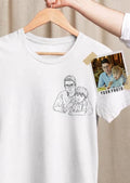 Fotografía en plano medio de una camiseta blanca de manga corta de la marca Trnd.Ink, colgada en un gancho de madera sostenido por una mano. En el lado izquierdo del pecho se muestra un estampado minimalista en estilo line art (retrato de trazos negros finos) que ilustra a un padre abrazando a su hijo pequeño. A la derecha de la prenda, fijada con cinta adhesiva a modo de referencia, aparece una fotografía real de un papá y su niño con el texto "YOUR PHOTO", destacando el proceso de personalización.