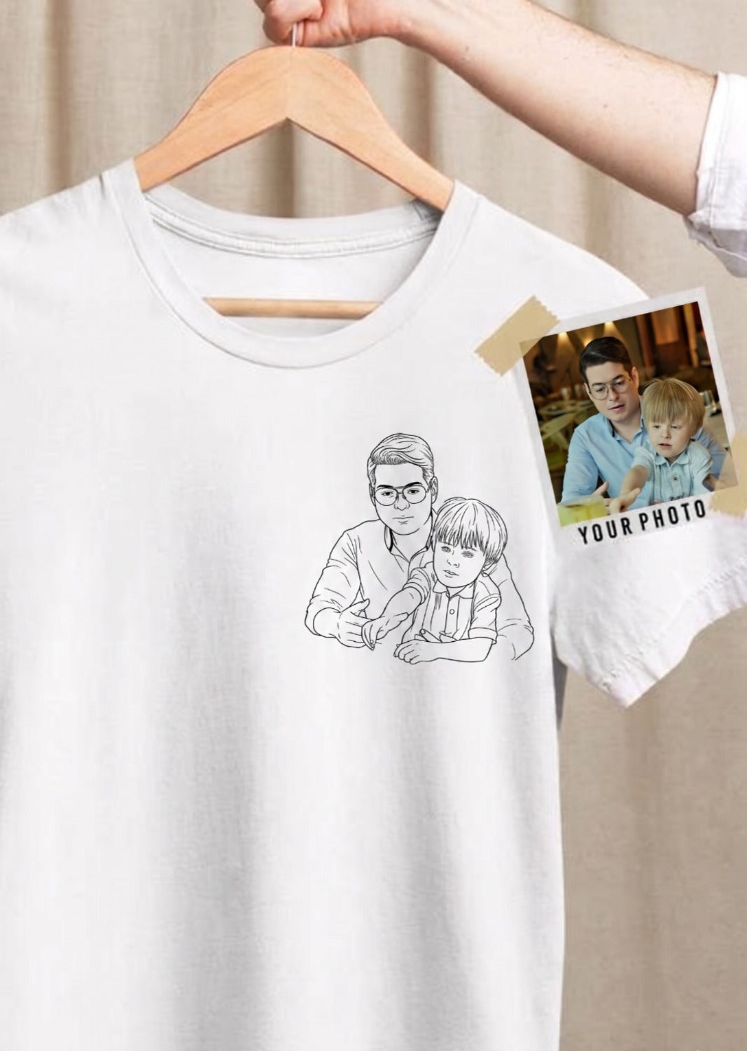 Fotografía en plano medio de una camiseta blanca de manga corta de la marca Trnd.Ink, colgada en un gancho de madera sostenido por una mano. En el lado izquierdo del pecho se muestra un estampado minimalista en estilo line art (retrato de trazos negros finos) que ilustra a un padre abrazando a su hijo pequeño. A la derecha de la prenda, fijada con cinta adhesiva a modo de referencia, aparece una fotografía real de un papá y su niño con el texto "YOUR PHOTO", destacando el proceso de personalización.