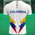 Vista posterior de una camiseta de fútbol blanca de la marca Trnd.Ink inspirada en la Selección Colombia. En la zona de la espalda resalta la palabra "COLOMBIA" estampada en letras grandes y mayúsculas de color azul oscuro. La parte inferior trasera de la prenda presenta un diseño geométrico abstracto con formas angulares en los colores de la bandera colombiana (amarillo, azul y rojo), mientras que en la parte superior central, justo debajo del cuello, se incorpora el logotipo de la marca alineado verticalm