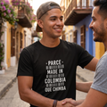 Hombre joven sonriendo con camiseta negra que dice "Parce Made in Colombia Que Chimba" en una calle colorida de estilo colonial.