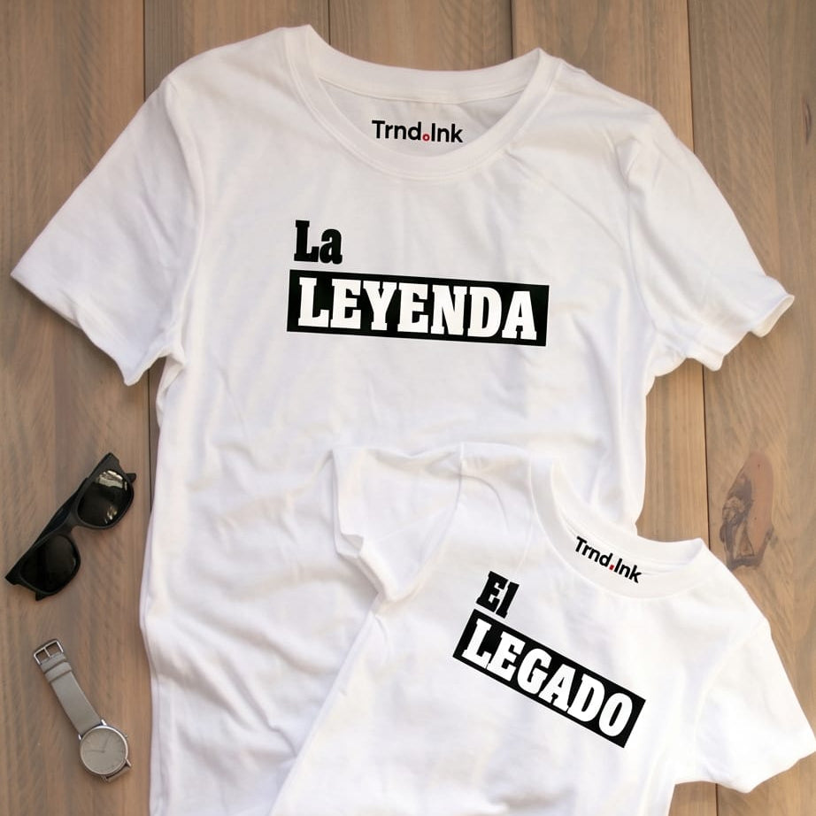Vista plana de dos camisetas blancas a juego para papá e hijo sobre un fondo de madera clara, con el logo de Trnd.Ink ubicado en la parte superior central de ambas prendas. La camiseta grande lleva el estampado "La Leyenda" y la pequeña "El Legado", decoradas a los lados con unas gafas de sol oscuras y un reloj de mano plateado.