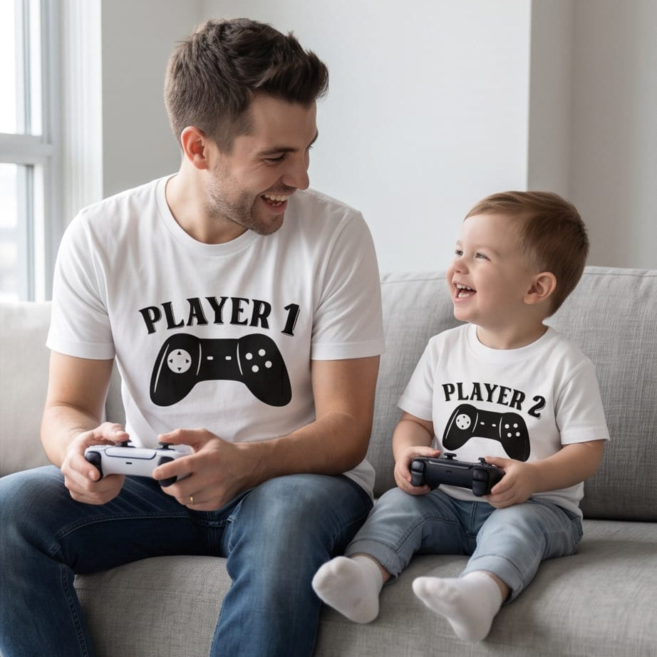 Fotografía de estilo de vida de un padre y su hijo pequeño sentados sonriendo en un sofá gris mientras juegan videojuegos con controles en las manos. Ambos visten camisetas blancas de la marca Trnd.Ink; la del papá lleva el estampado negro de un control clásico con el texto "PLAYER 1" y la del niño el texto "PLAYER 2" en un diseño minimalista.