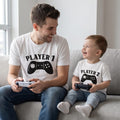 Fotografía de estilo de vida de un padre y su hijo pequeño sentados sonriendo en un sofá gris mientras juegan videojuegos con controles en las manos. Ambos visten camisetas blancas de la marca Trnd.Ink; la del papá lleva el estampado negro de un control clásico con el texto "PLAYER 1" y la del niño el texto "PLAYER 2" en un diseño minimalista.