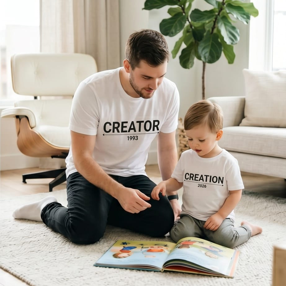 otografía de estilo de vida de un padre y su hijo pequeño sentados sobre una alfombra clara en una sala iluminada, mirando juntos un libro de cuentos abierto en el suelo. Ambos visten camisetas blancas a juego de la marca Trnd.Ink; la del papá lleva impreso el texto negro "CREATOR 1993" y la del niño "CREATION 2026" con un diseño minimalista. Al fondo se observa una planta de interiores y decoración moderna.