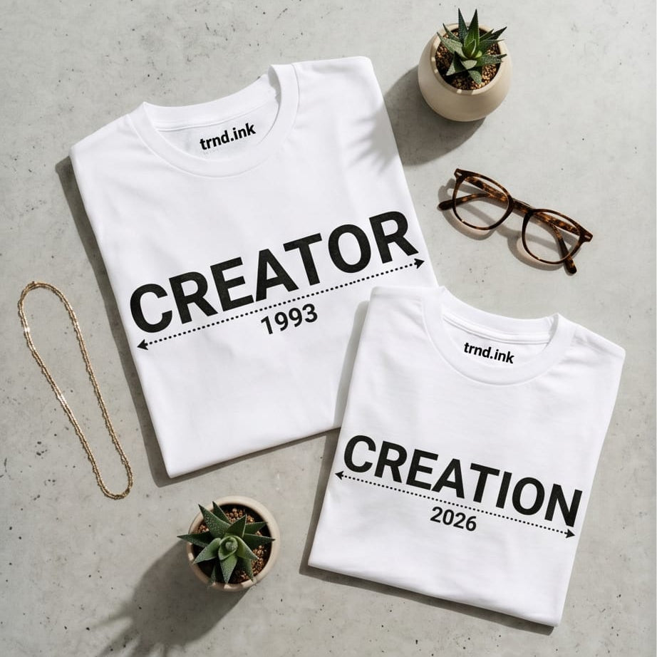 Composición flat lay de dos camisetas blancas dobladas a juego para papá e hijo sobre un fondo gris texturizado de la marca Trnd.Ink. La camiseta de adulto tiene el estampado negro "CREATOR" con una flecha y el año "1993", y la infantil lleva "CREATION" con el año "2026". El logo de Trnd.Ink está visible en la parte superior central de la etiqueta interna del cuello, rodeado de accesorios como gafas, una cadena dorada y plantas suculentas.
