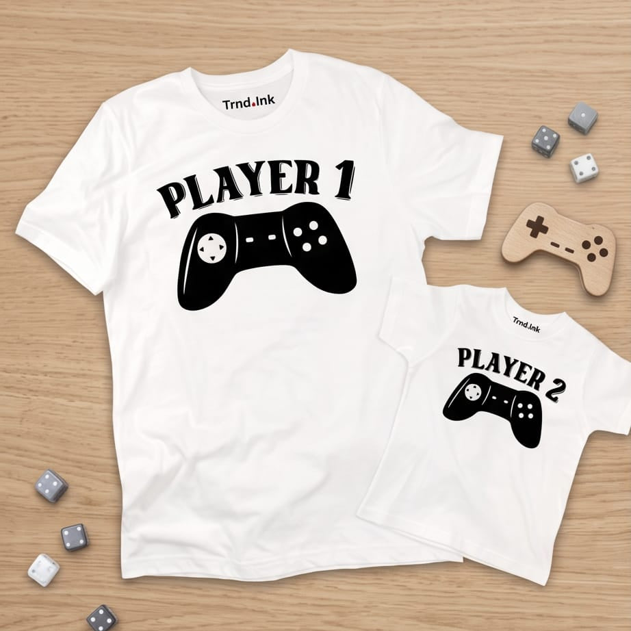 Vista plana estilo flat lay de dos camisetas blancas a juego para papá e hijo sobre un fondo de madera clara de la marca Trnd.Ink. La camiseta de adulto tiene el estampado negro "PLAYER 1" con un control de videojuegos clásico y la prenda infantil lleva "PLAYER 2"; ambas muestran el logo de Trnd.Ink ubicado en la parte superior central interna del cuello. La escena está decorada con pequeños dados grises y blancos y un control de madera.