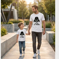 : Fotografía de estilo de vida de un padre y su hijo pequeño caminando y sonriendo de la mano por un sendero pavimentado al aire libre. Ambos visten camisetas blancas de manga corta de la marca Trnd.Ink; la del papá lleva un estampado negro con el casco de Darth Vader y la icónica frase "YO SOY TU PADRE", mientras que la del niño muestra el mismo casco con la frase "YO SOY TU HIJO".