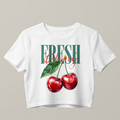 Vista frontal de un crop top blanco para mujer con un estampado gráfico central que muestra una ilustración detallada de dos cerezas rojas y hojas verdes, superpuestas con el texto "FRESH" en tipografía verde y "Cherry" en caligrafía roja, estilo streetwear minimalista de trnd.ink.