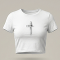 Crop top femenino de color blanco con ajuste ceñido, mostrando un diseño central de una cruz minimalista en trazo fino negro que integra la palabra "fé" en caligrafía elegante. La prenda se presenta sobre un fondo gris neutro con iluminación de estudio.