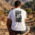 Un plano posterior de un hombre musculoso en cuclillas en un sendero de montaña rocoso y árido. El hombre está de espaldas a la cámara y lleva una camiseta blanca de manga corta y pantalones cortos grises. En la parte posterior de la camiseta hay un gráfico grande y centrado que muestra un casco de gladiador romano en tonos grises y plateados, con la palabra "VS" en letras blancas mayúsculas sobre el casco y las palabras "ME" en letras grises mayúsculas a ambos lados. El fondo es un paisaje montañoso con co