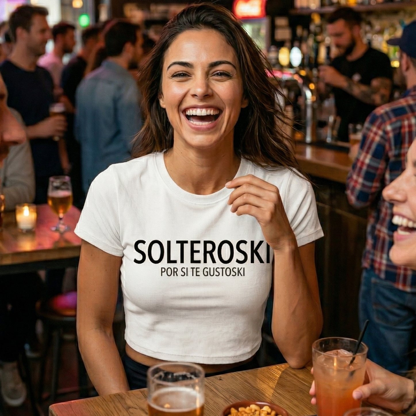 Modelo joven riendo en un bar luciendo una camiseta blanca tipo crop top con el texto SOLTEROSKI POR SI TE GUSTOSKI' en letras negras