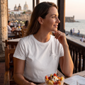 Una toma de ángulo medio de una mujer sonriente con cabello castaño sentada en un restaurante al aire libre en la azotea de Cartagena, Colombia. Ella lleva una camiseta corta blanca con dos colibríes negros dibujados en una línea continua estampados en el pecho. Su mano derecha descansa en su barbilla, mirando hacia la derecha. Frente a ella, sobre una mesa de madera, hay un tazón de cristal con ensalada de frutas frescas y yogur. El fondo muestra una vista panorámica de la ciudad vieja de Cartagena con sus
