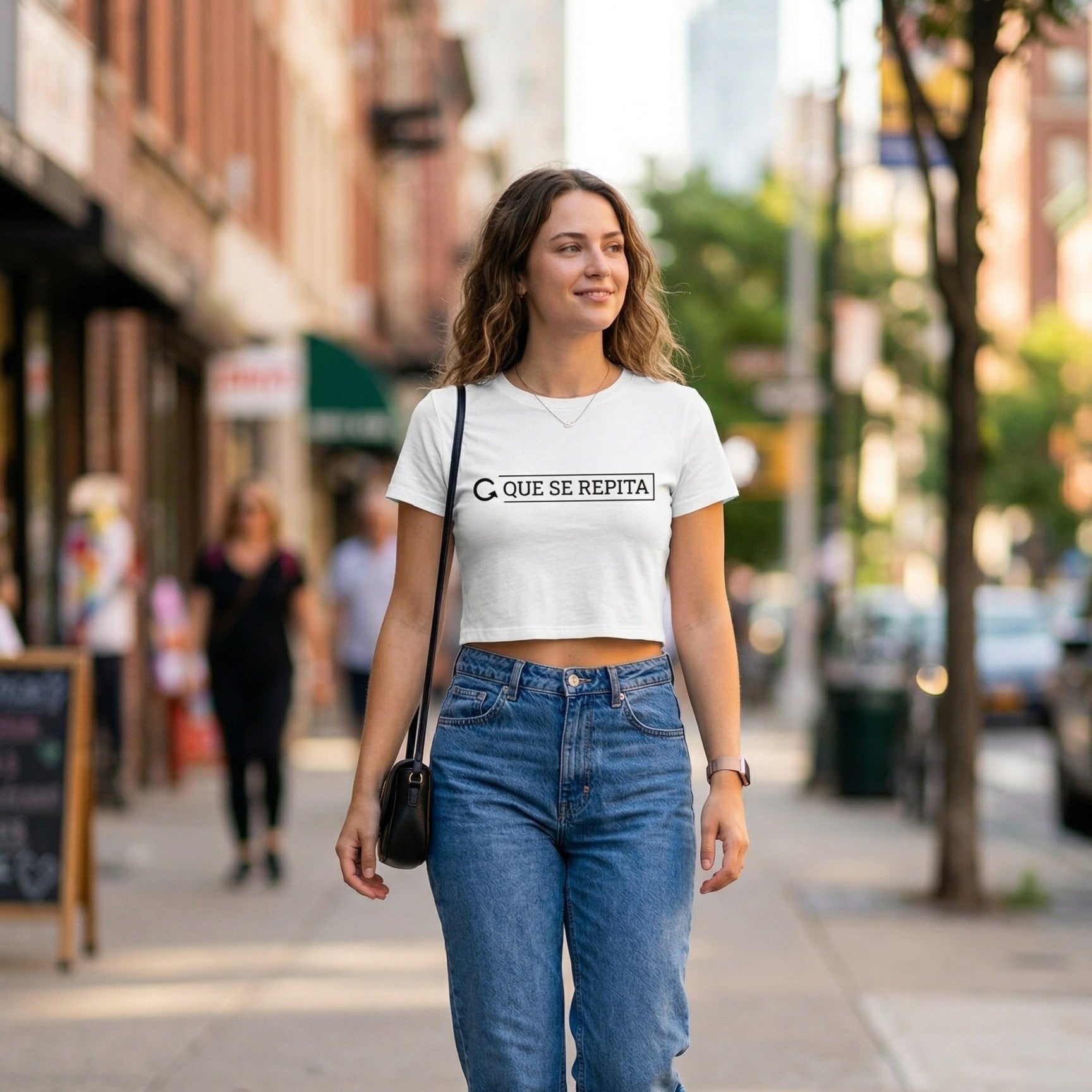 "Mujer joven caminando por una calle urbana con un outfit casual compuesto por un crop top blanco de Trnd.Ink con la frase 'QUE SE REPITA', jeans azules de tiro alto y un bolso pequeño negro. La imagen tiene un fondo de ciudad desenfocado con luz natural de día.