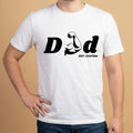 Mockup de un modelo masculino vistiendo una camiseta blanca de manga corta y jeans oscuros de la marca Trnd.Ink. En el pecho destaca un estampado gráfico central en tinta negra con la palabra "DAD", donde la letra 'A' está creativamente reemplazada por la silueta de un brazo musculoso flexionando el bíceps, acompañado abajo por el texto "EST. CUSTOM" en tipografía pequeña, todo sobre un fondo plano de color beige claro.