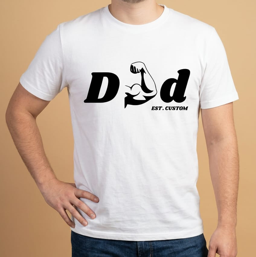 Mockup de un modelo masculino vistiendo una camiseta blanca de manga corta y jeans oscuros de la marca Trnd.Ink. En el pecho destaca un estampado gráfico central en tinta negra con la palabra "DAD", donde la letra 'A' está creativamente reemplazada por la silueta de un brazo musculoso flexionando el bíceps, acompañado abajo por el texto "EST. CUSTOM" en tipografía pequeña, todo sobre un fondo plano de color beige claro.