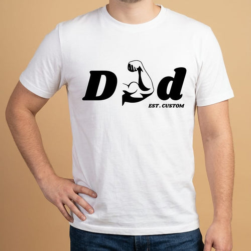 Mockup de un modelo masculino vistiendo una camiseta blanca de manga corta y jeans oscuros de la marca Trnd.Ink. En el pecho destaca un estampado gráfico central en tinta negra con la palabra "DAD", donde la letra 'A' está creativamente reemplazada por la silueta de un brazo musculoso flexionando el bíceps, acompañado abajo por el texto "EST. CUSTOM" en tipografía pequeña, todo sobre un fondo plano de color beige claro.