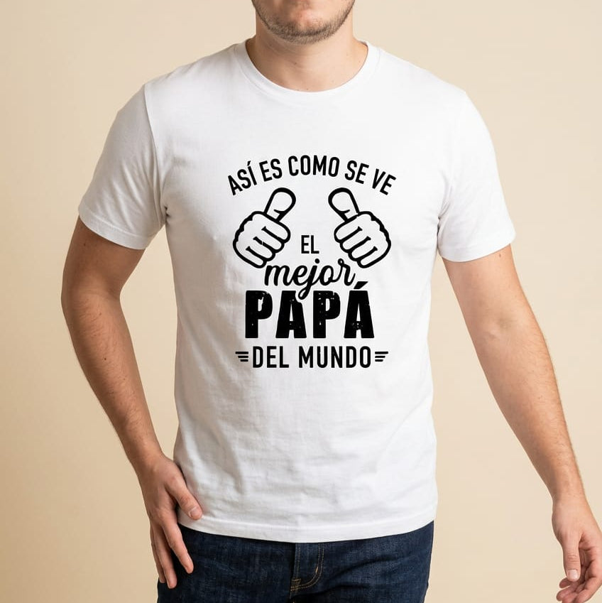 Mockup de un modelo masculino vistiendo una camiseta blanca de manga corta de la marca Trnd.Ink. En el pecho resalta un estampado gráfico central con tipografía negra de estilo minimalista y sarcástico que dice "ASÍ ES COMO SE VE EL mejor PAPÁ =DEL MUNDO=", acompañado por dos ilustraciones de manos con los pulgares arriba apuntando hacia el centro.