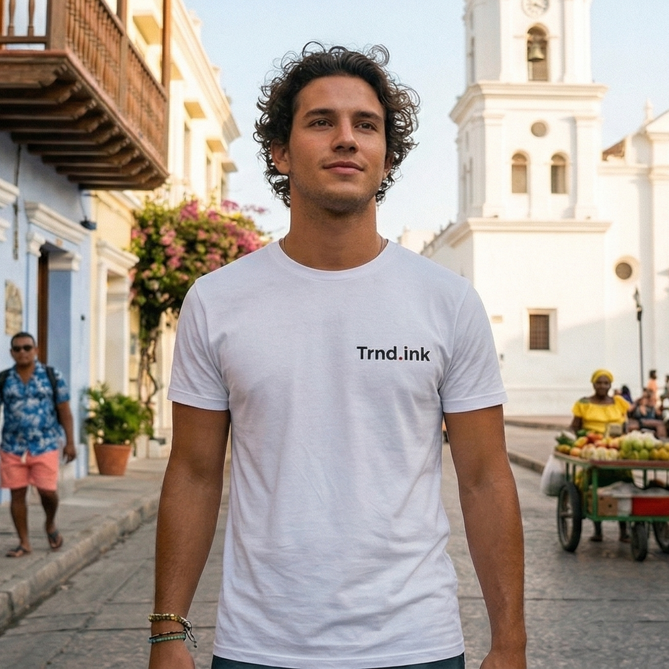 Hombre joven con cabello rizado vistiendo una camiseta blanca de la marca Trnd.ink con el logo en el pecho, caminando por una calle colonial con arquitectura antigua y una iglesia blanca al fondo bajo la luz del atardecer