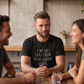 Un hombre barbudo sonriendo sutilmente en una mesa de un bar con amigos, vistiendo una camisa negra personalizada con la frase en español: 'A MI ME TRAJERON. YO NO QUERIA VENIR.