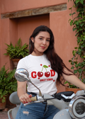 Mujer joven con cabello largo y jeans posando en una moto scooter gris, vistiendo una camiseta blanca de cuello redondo con el estampado 'GOOD' en letras rojas donde las 'O' son dos cerezas, y la frase 'Only Vibes' abajo en estilo urbano.