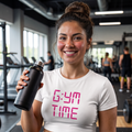 Camiseta deportiva blanca tipo crop top para mujer con el estampado "6:YM TIME" en letras rosadas estilo reloj digital. Modelo sonriendo en un gimnasio mientras sostiene un termo de agua negro.