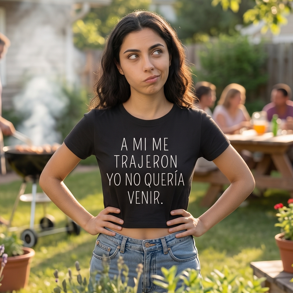 Camiseta negra tipo crop top para mujer con la frase de humor "A mí me trajeron yo no quería venir" en letras blancas. Modelo haciendo un gesto de sarcasmo en un jardín durante una reunión familiar o asado.