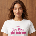 Camiseta blanca tipo crop top para mujer con la frase "Una Bad Bitch girl de lo' 90" estampada en letras color rosa. Modelo vistiendo la prenda con jeans de tiro alto sobre un fondo de madera.