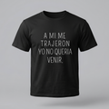 Una camiseta de algodón negra de manga corta con la frase en español 'A MI ME TRAJERON YO NO QUERIA VENIR.' impresa en la parte delantera en letras blancas finas y centradas. El fondo es de color gris claro.