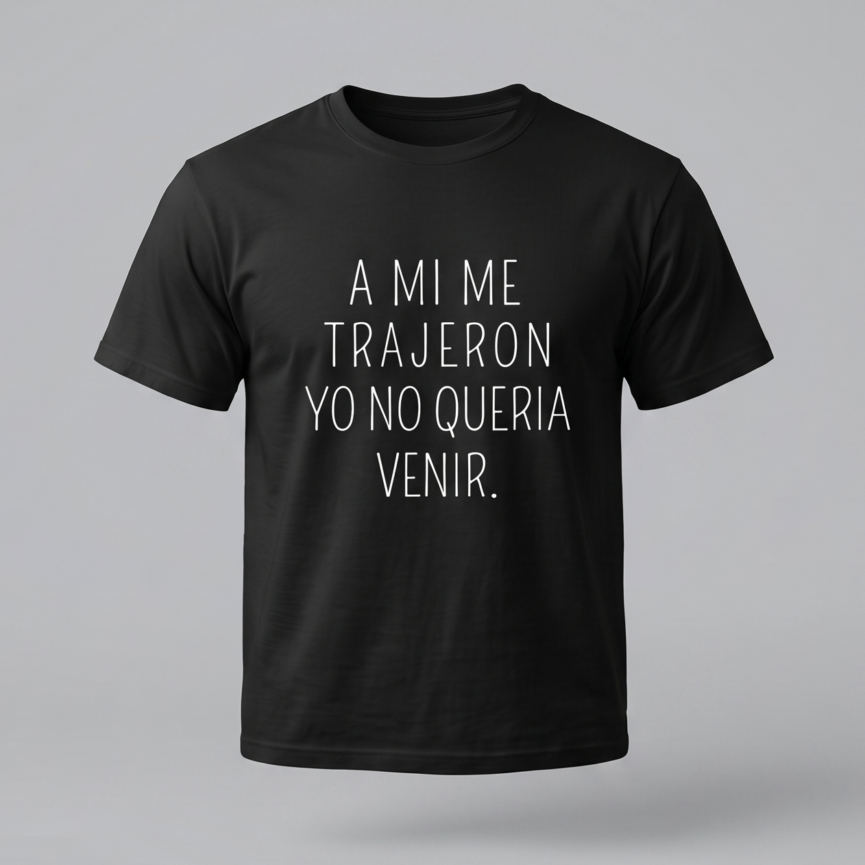 Una camiseta de algodón negra de manga corta con la frase en español 'A MI ME TRAJERON YO NO QUERIA VENIR.' impresa en la parte delantera en letras blancas finas y centradas. El fondo es de color gris claro.