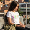Una mujer joven sentada en un banco de una estación de metro en Medellín, Colombia, usando una camiseta blanca con la frase '¿QUÉ MÁS PUES?' impresa en letras de colores. Está mirando su teléfono celular y tiene una bolsa de tela verde oliva a su lado. Al fondo se ve la ciudad y el metro elevado.