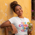 Camiseta blanca para mujer con la frase "Que Chimba de Vida" en letras coloridas de estilo urbano. Modelo sentada vistiendo la prenda y sosteniendo una bebida en un fondo amarillo.