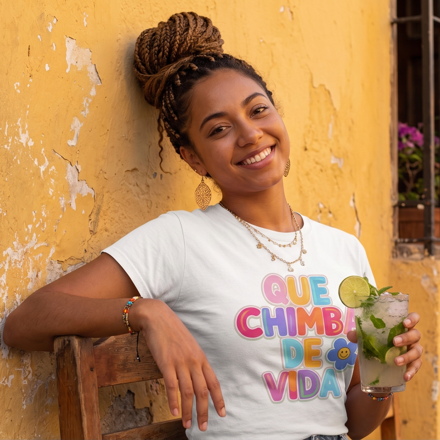 Camiseta blanca para mujer con la frase "Que Chimba de Vida" en letras coloridas de estilo urbano. Modelo sentada vistiendo la prenda y sosteniendo una bebida en un fondo amarillo.